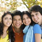 Four latino students -two girls and two boys- smiling directly at the camera, with a leafy background